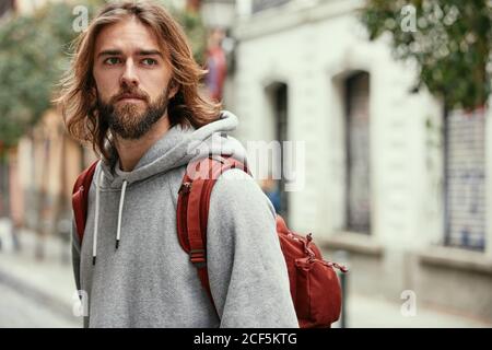 Jeune barbu beau homme en gris sweat à capuche avec sac à dos sérieusement en marchant dehors Banque D'Images