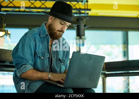 Jeune homme beau barbu avec chapeau noir assis et travaillant tenir le carnet sur des tours Banque D'Images