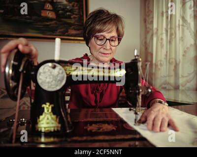 Femme senior en lunettes utilisant une machine à coudre rétro pour créer serviette en lin dans une chambre confortable à la maison Banque D'Images