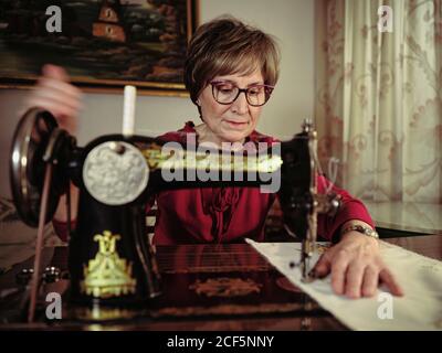 Femme senior en lunettes utilisant une machine à coudre rétro pour créer serviette en lin dans une chambre confortable à la maison Banque D'Images