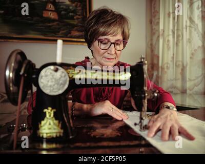 Femme senior en lunettes utilisant une machine à coudre rétro pour créer serviette en lin dans une chambre confortable à la maison Banque D'Images