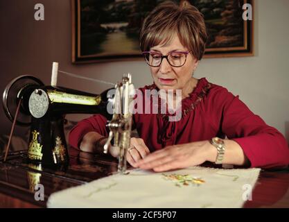 Femme senior en lunettes utilisant une machine à coudre rétro pour créer serviette en lin dans une chambre confortable à la maison Banque D'Images