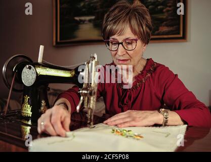 Femme senior en lunettes utilisant une machine à coudre rétro pour créer serviette en lin dans une chambre confortable à la maison Banque D'Images