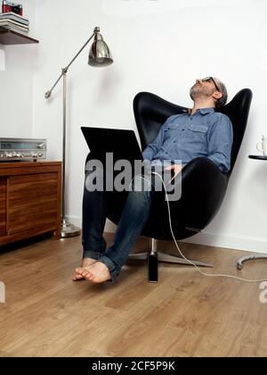 homme travaillant sur un ordinateur portable submergé sur un fauteuil à la maison Banque D'Images