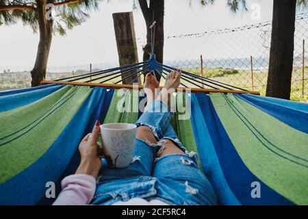 Rognez une femme dans un Jean allongé dans un hamac avec une tasse de café chaud et paysage tropical pendant les vacances d'été Banque D'Images