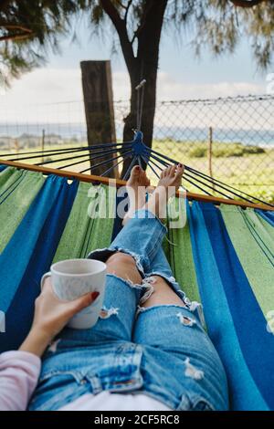 Rognez une femme dans un Jean allongé dans un hamac avec une tasse de café chaud et paysage tropical pendant les vacances d'été Banque D'Images