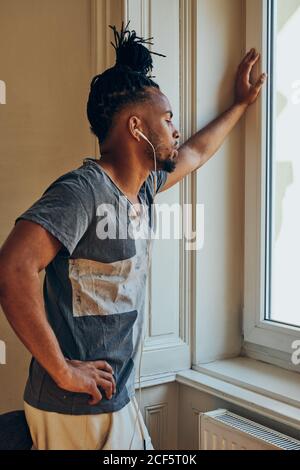 Vue latérale d'un homme ethnique sérieux avec une coiffure créative à l'écoute pour écouter de la musique et vous pencher sur le cadre de la fenêtre Banque D'Images