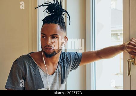 Vue latérale d'un homme ethnique sérieux avec une coiffure créative à l'écoute pour écouter de la musique et vous pencher sur le cadre de la fenêtre Banque D'Images