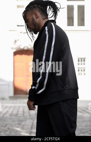 Jeune homme ethnique aux cheveux tressés portant un costume de sport noir en regardant vers le bas sur fond urbain Banque D'Images