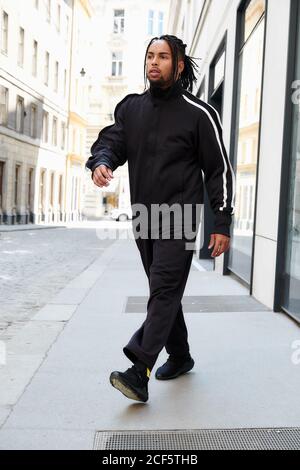 Homme afro-américain avec corneilles en costume de sport noir marchant sur fond urbain Banque D'Images