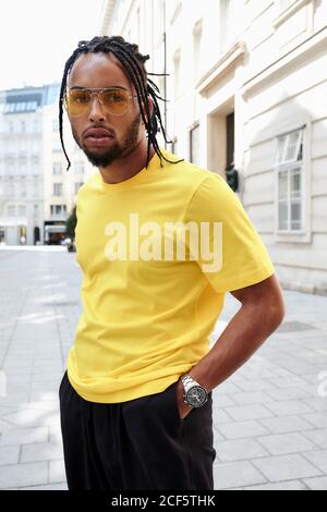 Jeune homme ethnique avec des cheveux tressés portant une chemise jaune et lunettes de soleil regardant l'appareil photo sur fond urbain Banque D'Images