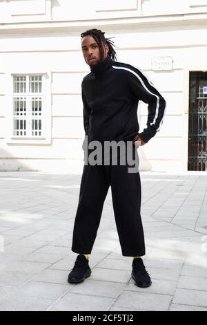 Jeune homme ethnique aux cheveux tressés portant un costume de sport noir regarder l'appareil photo sur fond urbain Banque D'Images