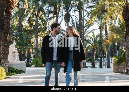 Joyeux jeune couple romantique en Jean et vestes noires tendance marcher dans une allée dans un parc avec des palmiers tropicaux et profiter vacances ensemble par temps ensoleillé Banque D'Images