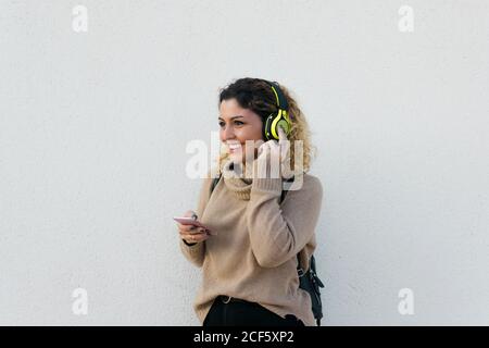 Jeune femme heureuse en beige chandail décontracté souriant en utilisant un casque et un téléphone portable avec un mur blanc sur fond Banque D'Images