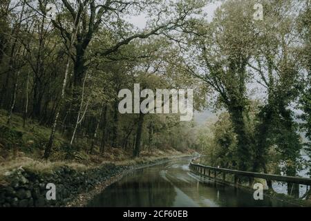 Paysage sombre avec une route asphaltée humide et sinueuse qui s'éloigne Forêt verte en terrain montagneux par temps couvert en Écosse campagne Banque D'Images