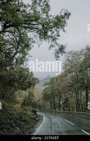 Paysage sombre avec une route asphaltée humide et sinueuse qui s'éloigne Forêt verte en terrain montagneux par temps couvert en Écosse campagne Banque D'Images