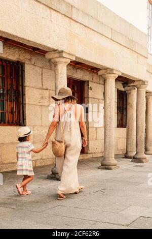 Vue arrière de la femme en chapeau et de l'enfant marchant à la main sur le trottoir le long de la vieille maison sur la colonne Banque D'Images