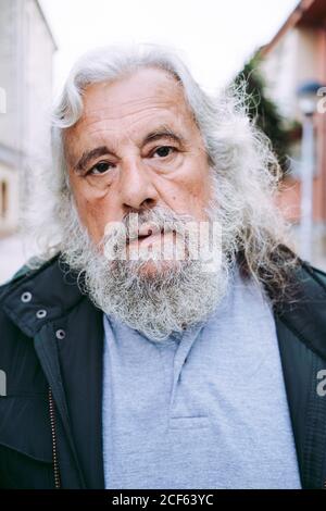 Sérieux senior homme retraité dans le port décontracté souriant à l'appareil photo en se tenant sur une rue floue en ville pendant la journée Banque D'Images