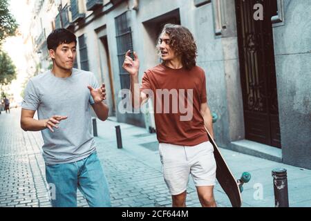 Sans souci intéressé multiethnique hommes dans des vêtements décontractés avec gestuelle de long-board et parler en flânant le long de la rue de la ville Banque D'Images