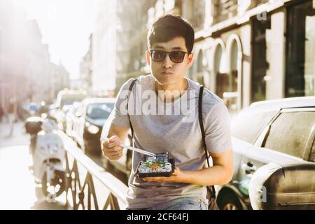 Homme asiatique élégant et ethnique en lunettes de soleil mangeant de la nourriture à emporter avec baguettes debout près d'un camion sur une rue ensoleillée Banque D'Images