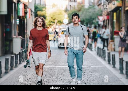 Sans souci intéressé multiethnique hommes dans des vêtements décontractés avec gestuelle de long-board et parler en flânant le long de la rue de la ville Banque D'Images