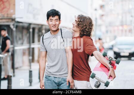 Sans souci intéressé multiethnique hommes dans des vêtements décontractés avec gestuelle de long-board et parler en flânant le long de la rue de la ville Banque D'Images