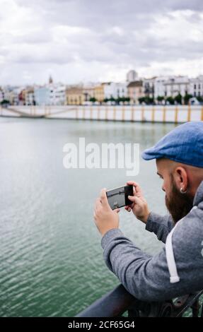Vue latérale de la taille basse à barbes pour homme, style décontracté prendre des photos de la rivière sur un téléphone portable tout en étant debout pont au quai Banque D'Images