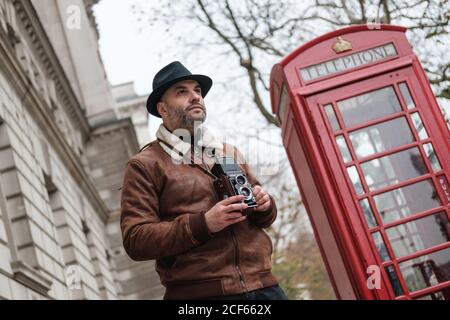 D'en dessous, un homme avec un appareil photo rétro regardant loin pendant situé sur la rue London, à proximité d'un téléphone rouge traditionnel Banque D'Images