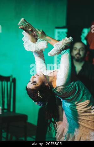 Danseuse hispanique adulte en robe rouffée prenant un fan de main tout en dansant avec les bras levés devant le musicien barbu pendant la performance Banque D'Images