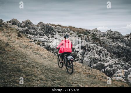 Vue arrière de la femme anonyme en casque noir et veste rouge vélo sur route rocheuse déserte le jour nuageux Banque D'Images