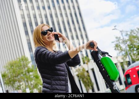 Vue latérale d'une femme chaude vêtue de lunettes de soleil parlant sur un téléphone portable tout en se déplaçant sur un scooter électrique Banque D'Images