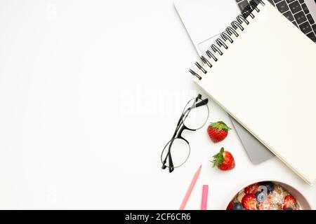 Vue de dessus du bloc-notes ouvert avec des pages vierges placées sur dessus du clavier du netbook près de la cuvette de petit déjeuner avec des baies Banque D'Images