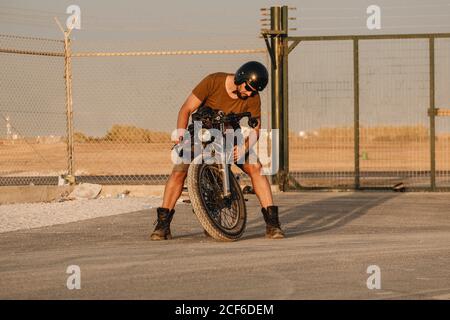 Homme concentré dans un casque et des lunettes de soleil fixant la moto sur le terrain Banque D'Images
