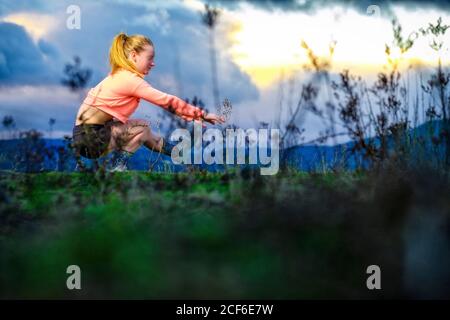 Vue latérale d'une femme puissante qui fait un squat sur une jambe contre le ciel nuageux au coucher du soleil pendant l'entraînement fonctionnel dans le champ Banque D'Images