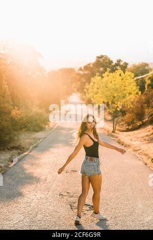 Vue arrière du contenu belle femme en jean short regardant sur l'épaule tout en marchant le long de la route de campagne pittoresque en contre-jour Banque D'Images