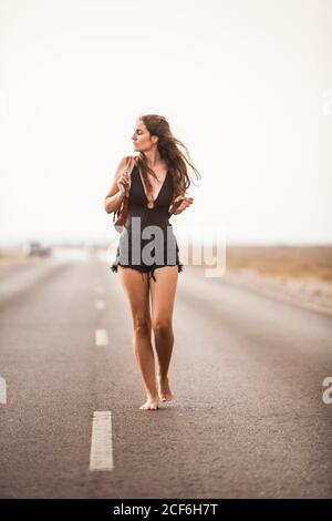 Belle jeune femme pieds nus souriant et marchant sur une route vide tenant sac à dos regarder loin Banque D'Images