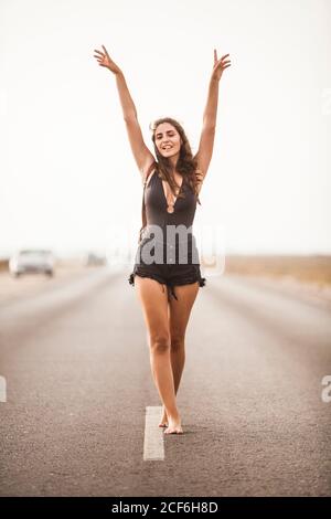 Belle jeune femme pieds nus souriant et marchant sur une route vide avec sac à dos levant les mains et regardant l'appareil photo Banque D'Images