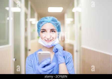 femme chirurgien debout dans le couloir tout en vérifiant les messages sur son smartphone, regarder l'appareil photo et sourire Banque D'Images