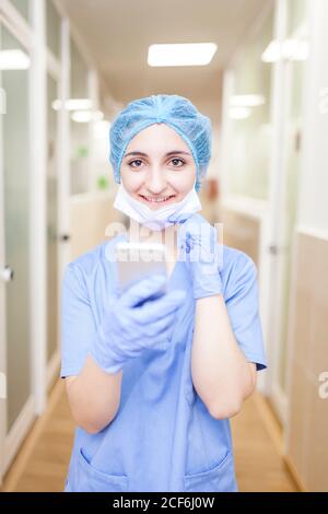 femme chirurgien debout dans le couloir tout en vérifiant les messages sur son smartphone, regarder l'appareil photo et sourire Banque D'Images