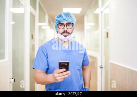 Un chirurgien masculin se tenant dans le couloir tout en vérifiant les messages sur son smartphone, regardez la caméra Banque D'Images