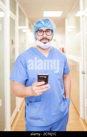 Un chirurgien masculin se tenant dans le couloir tout en vérifiant les messages sur son smartphone, regardez la caméra Banque D'Images