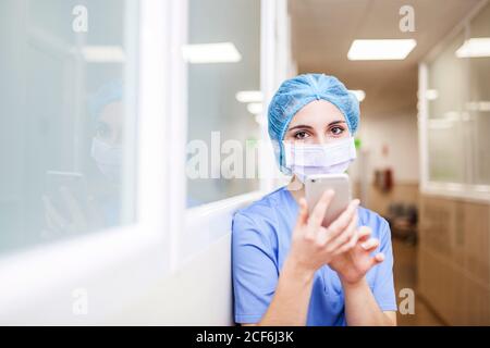 femme chirurgien debout dans le couloir tout en vérifiant les messages sur son smartphone, regarder l'appareil photo et sourire Banque D'Images