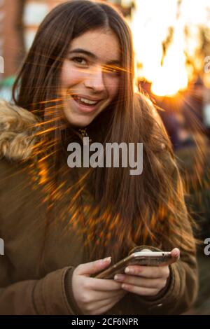 Jolie jeune femme dans une veste chaude souriant et regardant l'appareil photo tout en utilisant un smartphone dans la rue urbaine contre un soleil éclatant Banque D'Images