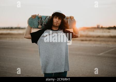 Un garçon adolescent taille basse élégant tenant une planche à roulettes derrière la tête et regarder la caméra sur une route vide au coucher du soleil Banque D'Images