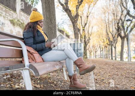 Vue latérale de la femme afro-américaine élégante pensive en chapeau jaune et veste chaude confortable assis sur un banc en bois et livre de lecture dans le parc d'automne Banque D'Images