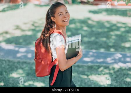 Bon étudiant. Jeune femme asiatique souriant à l'appareil photo, marchant sur le campus avec sac à dos, ordinateur portable, livre. Concept de retour à l'école Banque D'Images