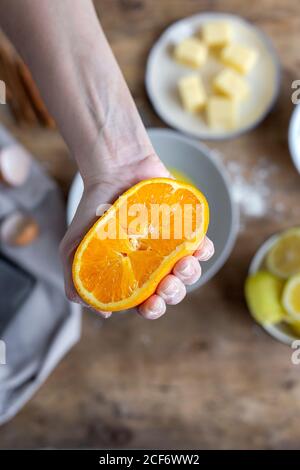 Du dessus de la main de récolte de femme méconnaissable recouverte de farine et montrant à l'appareil photo une moitié fraîche de couper l'orange sur le bol tout en préparant la pâte à la table Banque D'Images