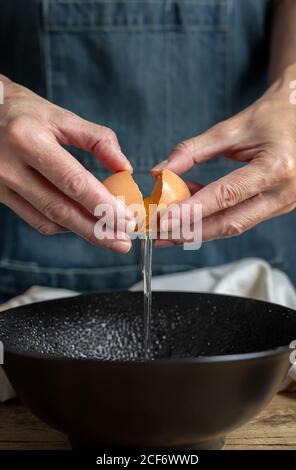 une femelle méconnaissable cassant un œuf de poulet frais dans un bol pendant la cuisson pâtisserie dans une table en bois avec des ingrédients frais Banque D'Images