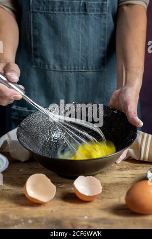 Récolte anonyme femme dans le tablier fouetter des oeufs dans le bol noir sur table en bois avec des ingrédients de citron , farine, beurre et cannelle bâtons pour le gâteau Banque D'Images