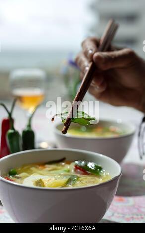 Par le dessus, une personne non reconnaissable et portant un bol de soupe orientale de nouilles ramen saines avec shiitake, épinards, carottes, œufs et piments sur la table du restaurant Banque D'Images